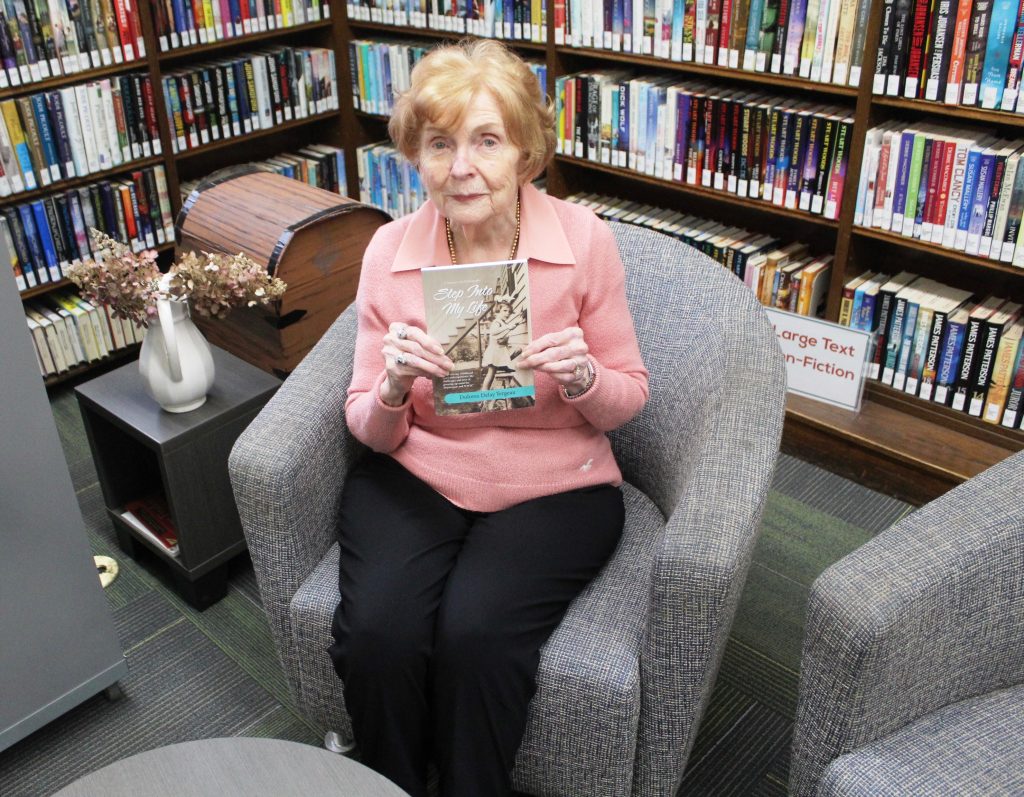 A photograph of Dolores Yergeau, seated in the library, holding a copy of her recent memoir called, "Step Into My Life."